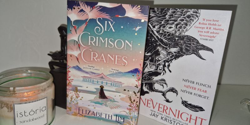 Two books, Six Crimson Cranes by Elizabeth Lim and Nevernight by Jay Kristoff) on a white shelf with a candle