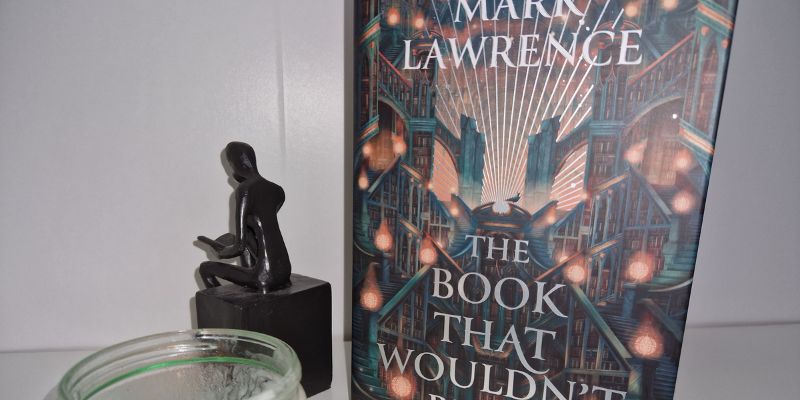 A book (The Book that Wouldn't Burn by Mark Lawrence) displayed on a white shelf with a bookend showing a girl reading a book and a candle from Istoria